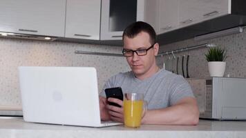 A young man during remote work from home writes data from a phone to a laptop while sitting at a table. video