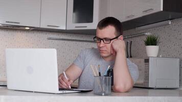 un joven masculino ingeniero en lentes es a un remoto trabajo a hogar y trabajos utilizando un ordenador portátil mientras sentado a un mesa. video