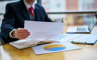 Mature businessman looking at laptop reading printed documents. Sitting near the window in office. Panoramic city view background. Business photo. photo