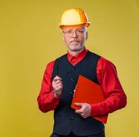 Engineer in a helmet on a yellow background. Senior man holding clipboard. photo