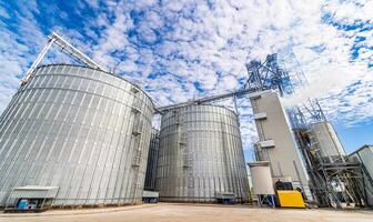 Agricultural silo, foreground sunflower plantations. Building exterior. Storage and drying of grains, wheat, corn, soy, sunflower against the blue sky with white clouds photo