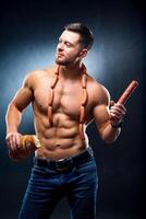 Bodybuilder with naked torso. Smoked sausage in one hand, sausages on a neck and fresh bread in other. Cropped photo. Lifestyle concept. Studio photo. Hungry sportsman. photo