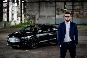 Stylish handsome man with sunglasses in fashionable clothes standing with the luxury black car on the background. Closeup photo