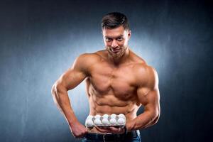 Handsome young sportsman is holding a packaging with eggs, healthy food protein, gray background. Naked torso, healthy life concept. photo