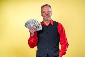 Portrait of happy and white teeth smile senior old business man holding money in hands, dressed in red shirt, isolated on yellow background. Human emotions and facial expressions photo