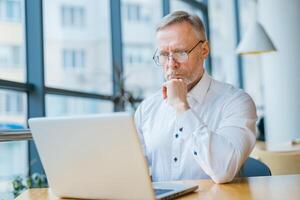Thoughtful businessman is reading important information on laptop. Photo in front of the window with city view. Business process.