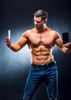 Bodybuilder sportsman holding sport bar and chocolate . Choosing between healthy and harmful food. Naked torso. Grey background. Vertical photo. Closeup photo