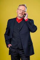 Portrait of a senior businessman standing against yellow background hand in pocket. Talking over phone. photo