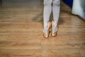 piernas de un pequeño bailarina en un oro ballet Zapatos y blanco calcetines posando punta del pie en ballet salón en el piso. de cerca. foto