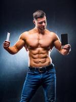 Bodybuilder sportsman holding sport bar and chocolate . Choosing between healthy and harmful food. Naked torso. Grey background. Vertical photo. Closeup photo