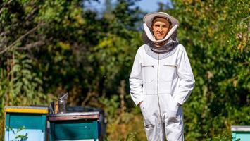 Horizontal portrait of a beekeeper surrounded with hives and trees at the apiary. Man with hands in pockets in white uniform. photo