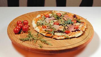 Appetizing pizza rotating and lying on a wooden board with cherry tomatoes. Cooked pizza with prosciutto, arugula and tomatoes. Delicious fast food. Close up video