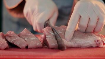 A chef in gloves cuts meat into pieces with a knife. Preparation of meat for cooking steaks. Close up video