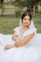 Portrait. The brunette bride, smiling sincerely, sits on the ground in her unfolded dress, posing. Wedding ceremony. Sunny day. Beautiful hair and makeup photo