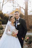 elegante, joven novio y hermosa novia en un largo blanco vestir y un largo velo con un ramo de flores en su manos, abrazando en el parque en el otoño naturaleza. Boda retrato de recién casados. foto