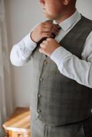a young man in a white shirt stands by the window in the room and puts on a tie. The groom gets dressed in the morning and prepares for the wedding. photo