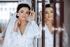 In the morning, an incredibly beautiful and luxurious bride, dressed in a white robe, poses by the mirror in her bedroom. MORNING of the bride with a luxurious hairstyle and natural makeup. photo