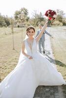 Wedding photo. A brunette bride in a voluminous lace dress holds a bouquet and walks in the garden in front of the groom, smiling sincerely. Gorgeous hair and makeup. Fashion and style. celebration. photo