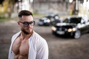 Handsome man is standing in unbuttoned white shirt near broken concrete wall. Abandoned storehouse and car on the background photo