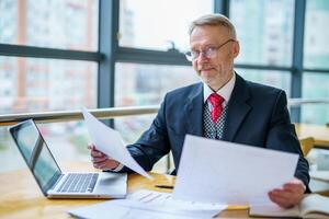 Mature businessman looking at laptop reading printed documents. Sitting near the window in office. Panoramic city view background. Business photo. photo