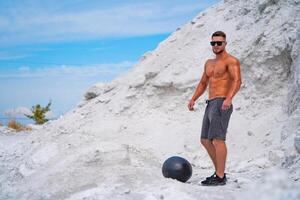 Muscle athlete strongman in sunglasses stands near black fitball. White sandy mountains or in a quarry. Sunny day. White landscape. photo