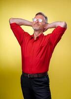 Handsome middle age senior man wearing a red shirt over isolated yellow background. Relaxing and stretching with arms and hands behind head and neck. photo