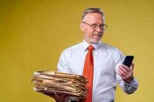 Studio shot of senior businessman looking at phone holding pile of papers. Yellow background photo