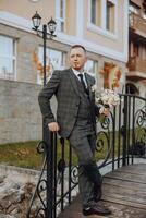 Portrait of a handsome young groom on an autumn day outdoors in an elegant suit and holding a wedding bouquet of flowers. Outdoor photo. Handsome man in a business suit. photo