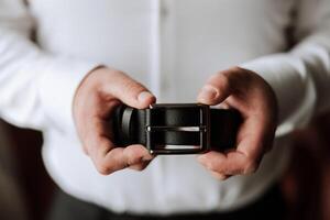 Close-up of a man holding a black leather belt. A man in a white shirt, without a face, holds a black leather belt in his hands. The morning of the groom at the wedding. photo