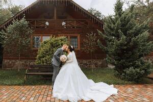 Handsome groom and charming bride stay together near modern wooden house in park. Wide-angle photo, place for text photo