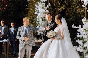 A master of ceremonies gives a speech while a bride and groom hold hands near a white floral arch photo