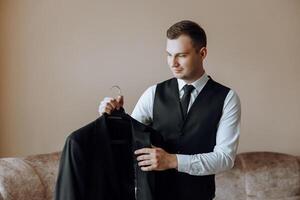 An elegant groom in a white shirt and a stylish tie is holding a jacket on a hanger. Details of the wedding morning in style. photo