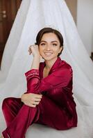 morning portrait of a luxurious and beautiful bride in a hotel in a luxurious room in red pajamas sitting and posing next to her wedding dress. photo