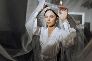 portrait of an incredibly beautiful girl bride in a white robe in the bedroom, the bride poses holding her veil in her hands and covers herself with it. photo