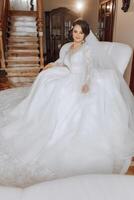 An incredibly beautiful bride sits on a bed in her bedroom. Bride morning. A gorgeous bride in a beautiful dress is sitting on a bed in a bright room . photo