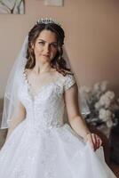 fashion photo of a beautiful bride with dark hair in an elegant wedding dress and stunning makeup in the room on the morning of the wedding. The bride is preparing for the wedding