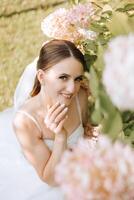 Portrait. The brunette bride, smiling sincerely, sits on the ground in her unfolded dress, posing near the flowers. Wedding ceremony. Sunny day. Beautiful hair and makeup photo