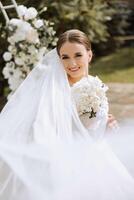 A brunette bride in a white dress holds a bouquet, poses. The veil is thrown into the air. Beautiful hair and makeup. Wedding photo session in nature.