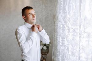 un hombre en un blanco camisa soportes por el ventana en el habitación y sujeta el botones en su collar y mangas reloj en mano. elegante negocio retrato de un hombre, de cerca foto. el novio es preparando. foto