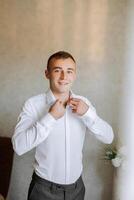 un hombre en un blanco camisa soportes por el ventana en el habitación y sujeta el botones en su collar y mangas reloj en mano. elegante negocio retrato de un hombre, de cerca foto. el novio es preparando. foto