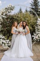 Wedding photography. The bride in a wedding dress and her friends in white dresses pose with bouquets near a white arch. photo