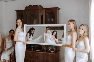 Bridesmaids rejoice in the morning, helping to fasten the buttons on the wedding dress and prepare for the wedding ceremony. They take pictures, smile, help the bride with her shoes. photo