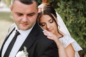 encantador y elegante recién casados son abrazando y sonriente en contra el antecedentes de otoño naturaleza en un hermosa jardín. un increíblemente hermosa joven novia inclinado en contra el hombro de su amado novio foto