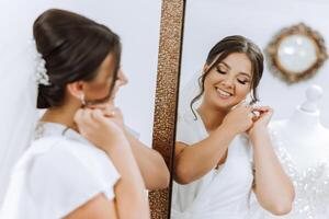 In the morning, an incredibly beautiful and luxurious bride, dressed in a white robe, poses by the mirror in her bedroom. MORNING of the bride with a luxurious hairstyle and natural makeup. photo