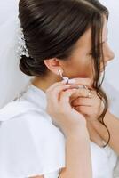 Beautiful young bride puts on earrings before wedding ceremony at home. A bride in a white robe puts on earrings in her room in the morning. photo