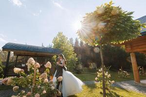 elegante novio sostiene hermosa novia en largo blanco vestir en su brazos y bailes en verde césped en otoño parque, bosque debajo luz de sol. Boda fotografía, retrato de sonriente recién casados. foto