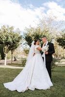 Stylish, young groom and beautiful bride in a long white dress and a long veil with a bouquet in their hands, hugging in the park in the autumn nature. Wedding portrait of newlyweds. photo