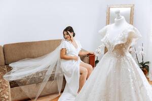 un increíblemente hermosa novia en el Mañana en un blanco túnica y un largo velo se sienta en el cama en su dormitorio. el novia poses en el Mañana antes de el Boda ceremonia. foto