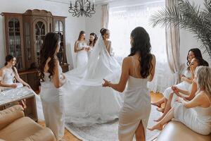 Bridesmaids rejoice in the morning, helping to fasten the buttons on the wedding dress and prepare for the wedding ceremony. They take pictures, smile, help the bride with her shoes. photo