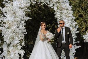 A stylish groom in a black suit and a cute bride in a white lace dress stand in a round arch photo